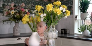 På bilden syns ett barn med en vit vas med gula konstgjorda påskliljor i ett kök. Bredvid barnet står en större vas med gula och vita blommor. I bakgrunden finns fler blommor i vaser, vita kaklade väggar och en blommigt mönstrad tapet.